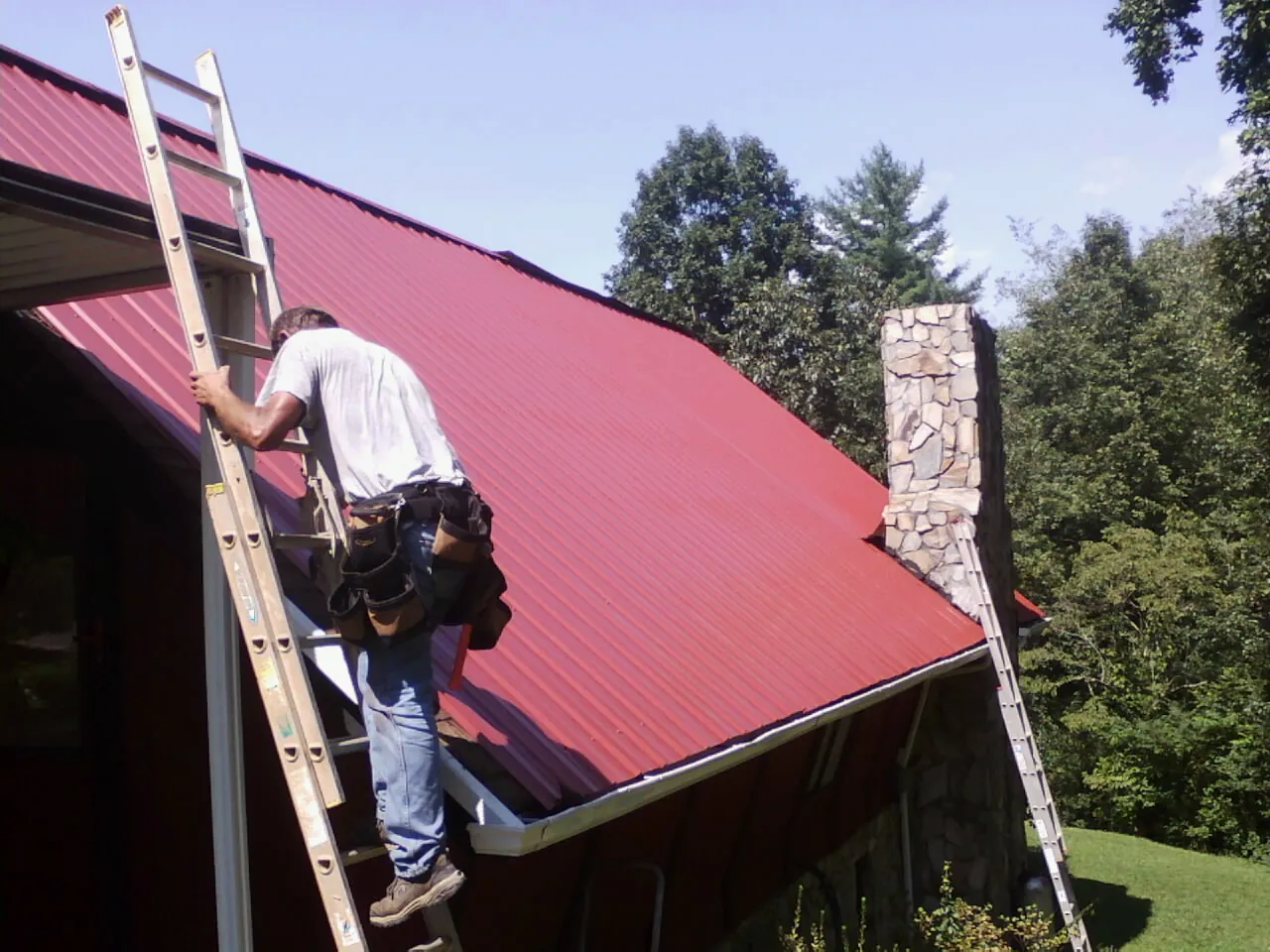 Roofer on a ladder beside a red metal roof and stone chimney — Soffit & Fascia Repair by Roofing Brothers in North Kingstown